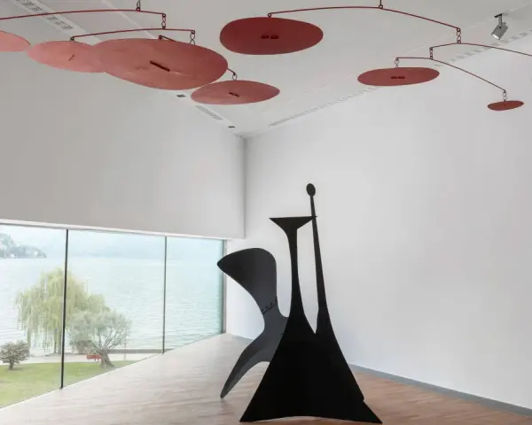 Un mobile Calder rouge flotte au-dessus d'une sculpture noire monumentale, dans une salle d'exposition lumineuse avec vue sur un lac.