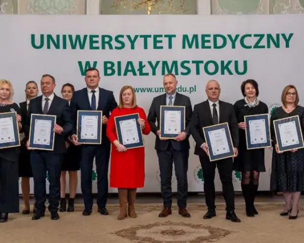 Grupa osób w garniturach i sukienkach pozuje do zdjęcia z dyplomami przed banerem Uniwersytetu Medycznego w Białymstoku.