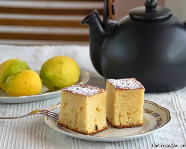 Dos porciones de bizcocho de limón espolvoreadas con azúcar glas, junto a limones frescos y una tetera negra.