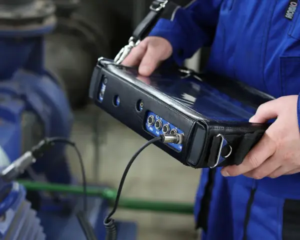Technik w niebieskim kombinezonie wykonuje pomiary drgań maszyny przemysłowej za pomocą przenośnego urządzenia.
