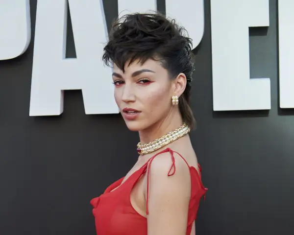 Mujer con un llamativo mullet largo, maquillaje rojo y vestido escarlata, posando ante un fondo con letras blancas.