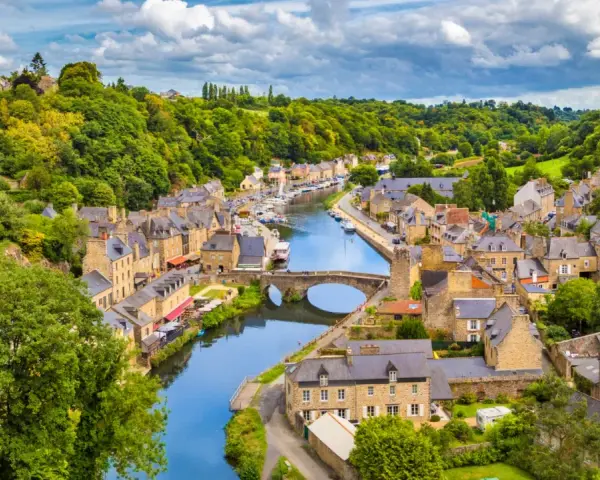 A picturesque view of Dinan, France, with its charming stone buildings lining a winding river, a stone bridge, and lush green hills under a cloudy sky.