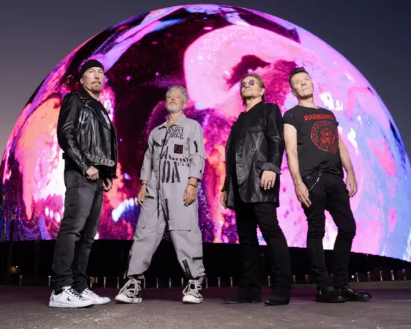 U2 w nowej płycie. Czterech członków zespołu stoi przed gigantycznym, kolorowym ekranem.
