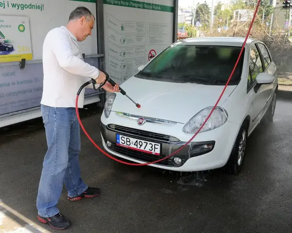 Mężczyzna myje białe auto na myjni. Na tablicy informacyjnej widnieje napis "Możliwość otrzymania faktury", sugerujący, że na myjni można płacić kartą.