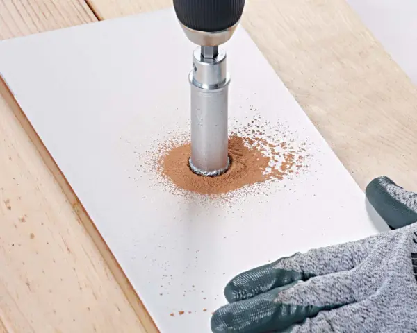 Une perceuse avec une couronne diamantée réalise une découpe carrelage, créant un nuage de poussière fine sur une plaque blanche.