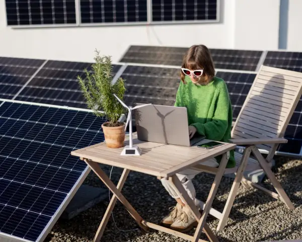 Dziewczyna w okularach pracuje na laptopie przy stole, otoczona panelami słonecznymi. Obok stoi model wiatraka, symbolizujący energię oze.