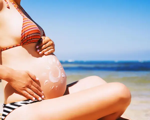 Ciężarna kobieta na plaży, z kremem przeciwsłonecznym na brzuchu w kształcie słońca. Bezpieczne opalanie natryskowe w ciąży.