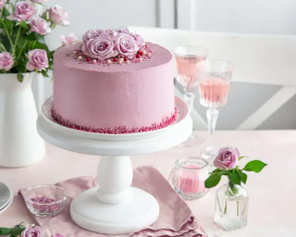 Różowy tort dla mamy, ozdobiony kwiatami i posypką. Obok kieliszki z różowym napojem i bukiet róż.