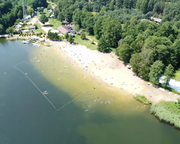 Słoneczny dzień nad jeziorem Rogoźnik. Ludzie kąpią się i opalają na piaszczystej plaży, otoczonej zielonym lasem.