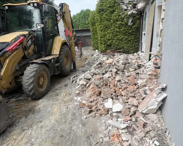 Żółta koparko-ładowarka pracuje przy wyburzeniu domu. Gruz i cegły leżą na ziemi. Koszt wyburzenia domu jest wysoki.
