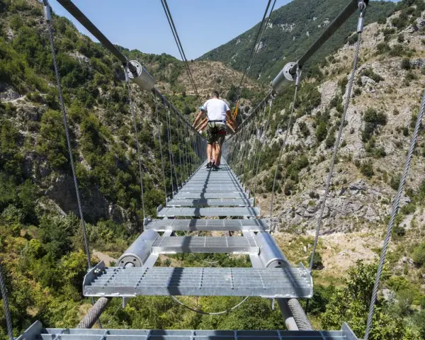 Un ponte tibetano in Puglia attraversa una gola verdeggiante, con persone che camminano su passerelle metalliche sospese tra montagne.