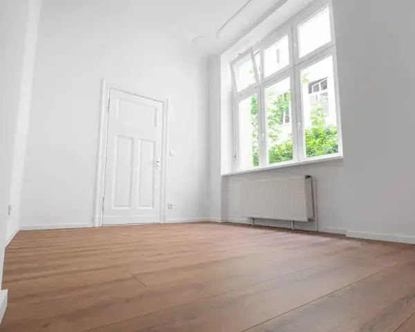 Puste, jasne pomieszczenie z białymi ścianami, drzwiami i oknem. Drewniana podłoga i grzejnik sugerują, że to gotowe do zamieszkania mieszkanie socjalne.