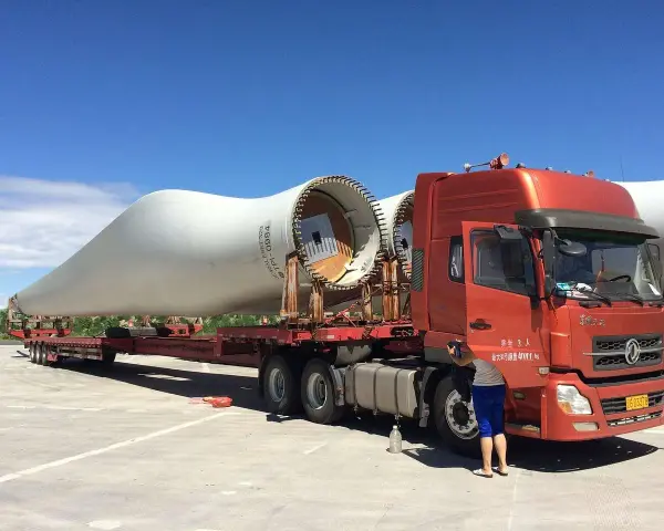Pomarańczowy tir przewozi gigantyczne łopaty turbin wiatrowych. Kierowca przygotowuje się do dalszej podróży.