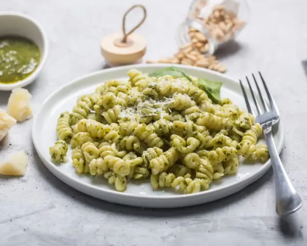 Pyszny makaron z pesto ze słoika i śmietaną, posypany parmezanem. Obok miseczka z pesto i kawałki sera.