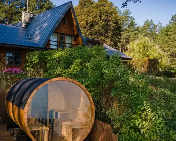 Drewniany domek nad jeziorem z beczką sauny w otoczeniu zieleni.