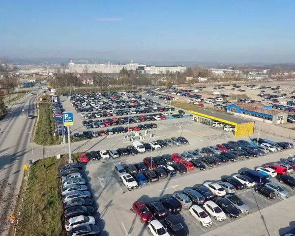Duży parking przy krakow katowice airport. Wiele samochodów zaparkowanych w rzędach, w tle budynki lotniska.