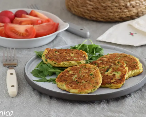 Tortitas de calabacín doradas y esponjosas, servidas sobre rúcula fresca. Acompañadas de una ensalada de tomate y rábano.