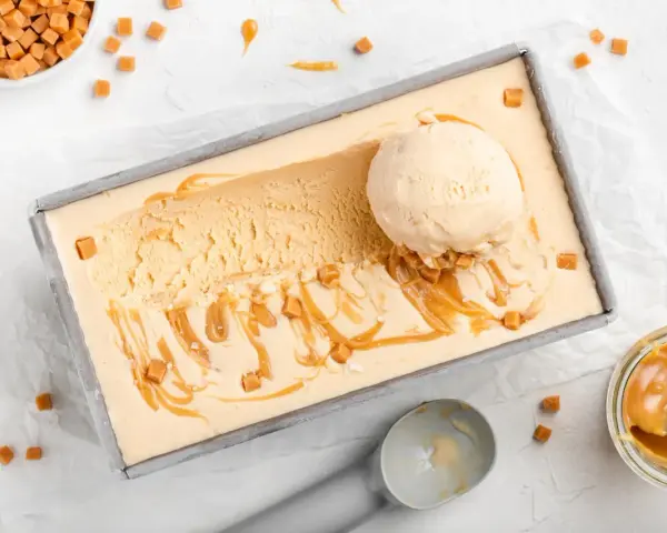 Ein Blech mit cremigem, hellbraunem Salted Caramel Eis, garniert mit Karamellsauce und kleinen Karamellwürfeln. Eine Eiskugel liegt darauf.