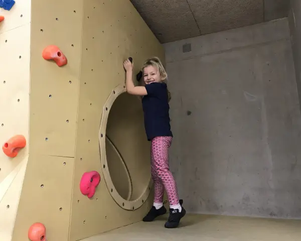 Ein fröhliches Mädchen klettert in einer Kletterhalle für Kinder. Sie lächelt und hält sich an einem Griff fest.
