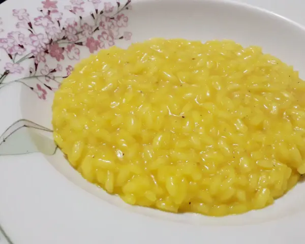 Un cremoso risotto allo zafferano, servito in un piatto bianco decorato con fiori rosa e rami verdi.