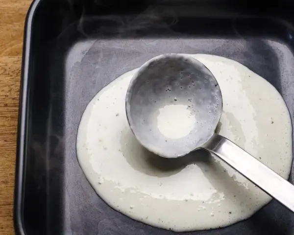Eine Schöpfkelle verteilt cremige Bechamel-Sauce in einer schwarzen Auflaufform.