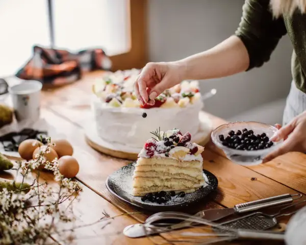 Manos expertas decoran un pastel en una mesa de reposteria y pasteleria, con frutas frescas y arándanos.