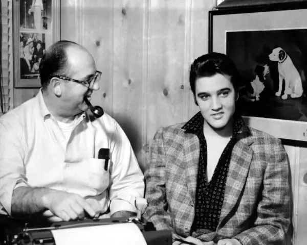 Elvis Presley und Colonel Parker, sein Manager, sitzen nebeneinander. Parker raucht Pfeife, Elvis blickt in die Kamera.