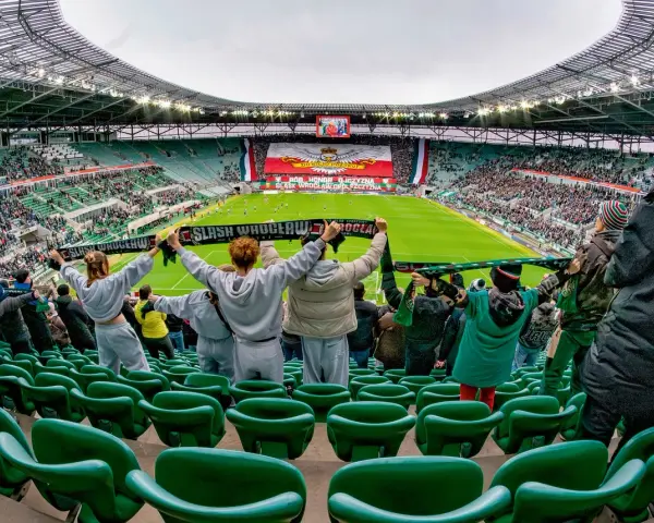 Gwardia Opole kibicuje na stadionie, trzymając szaliki. Na boisku trwa mecz, a trybuny wypełniają fani.