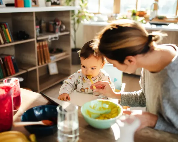 Mama karmi łyżeczką 6 miesięczne dziecko. Na stole są owoce i napoje.