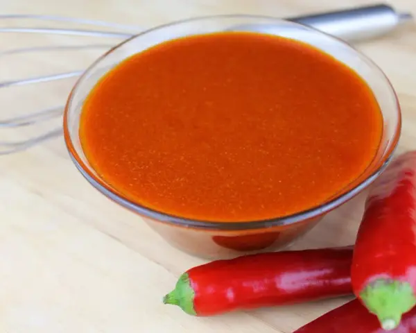 Schüssel mit leuchtend orangeroter Buffalo Sauce, daneben rote Chilis und ein Schneebesen. Perfekt für Flügel!