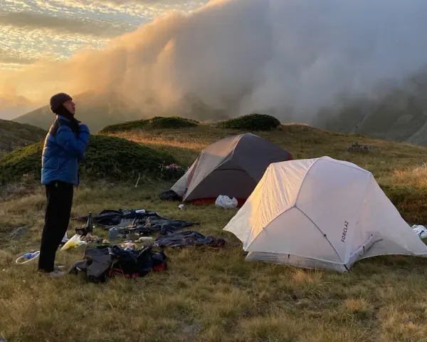 Une personne se tient près de tentes dans une aire de bivouac, profitant du lever du soleil sur les montagnes.