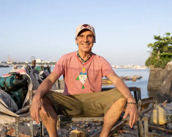 Le chanteur Mano Negra, souriant, assis sur un banc rouillé, avec un collier coloré et un t-shirt à motif poisson.