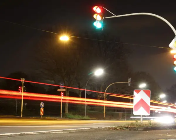 Nachts an einer Kreuzung mit roten Lichtspuren von Autos. Ampeln zeigen Grün und Rot. Ein Vorfahrtsschild und ein Richtungspfeil sind zu sehen. 132 bkat.