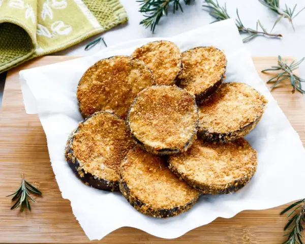 Cotolette di melanzane dorate e croccanti, servite su carta bianca con rametti di rosmarino.