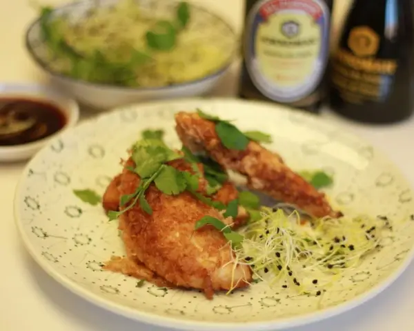 Crujiente solomillo de pollo empanado, adornado con cilantro fresco y brotes, servido con salsa y un cuenco de ensalada.