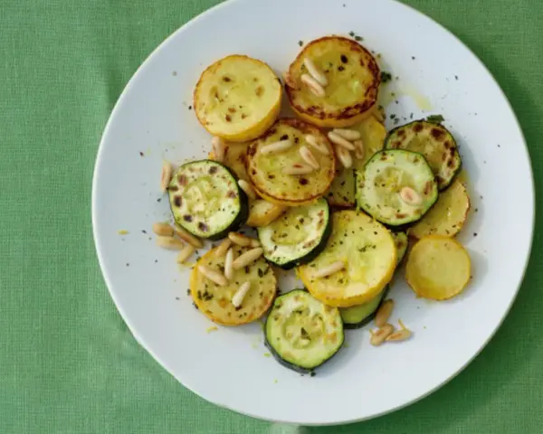 Gegrillte gelbe Zucchini-Scheiben mit Pinienkernen und Kräutern auf einem weißen Teller.