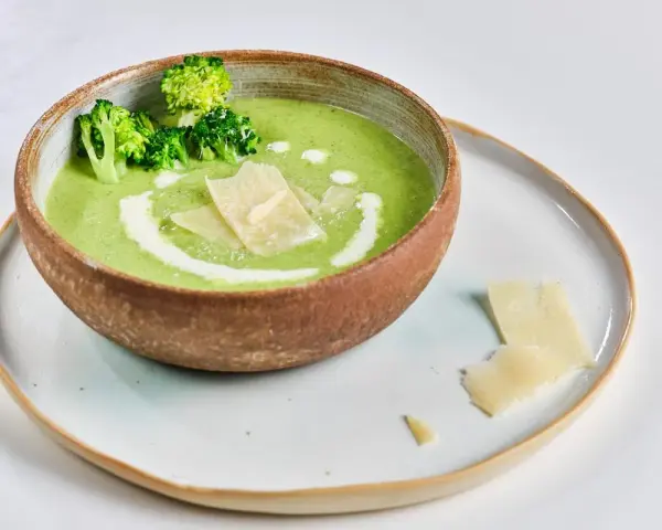 Crema de brócoli servida en un cuenco rústico, decorada con floretes de brócoli, un remolino de nata y láminas de queso parmesano.
