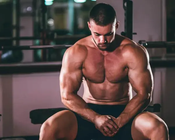 Un homme musclé, concentré, s'entraîne pour prendre du muscle rapidement. Il est assis dans une salle de sport, le regard baissé.