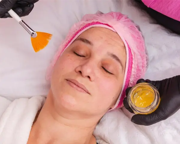 Kobieta w czepku leży z zamkniętymi oczami, a kosmetyczka przygotowuje maseczkę z miodu na twarz, używając pędzelka.