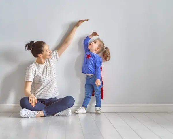 Mama mierzy wzrost córeczki w stroju superbohaterki. To właśnie wtedy dzieci rosną najszybciej, zaskakując nas każdego dnia.