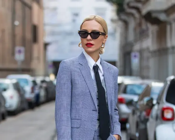 Estilo tomboy que es: mujer con traje a cuadros, camisa blanca y corbata negra, gafas de sol y labios rojos, posando en la calle.