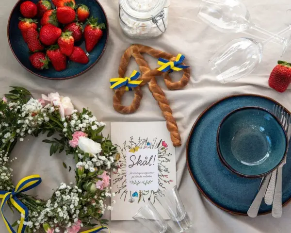 En midsommardukning med jordgubbar, blomsterkrans, en pretzel med svenska flaggor och en "Skål!"-bok. Ett personligt ljus för firandet.