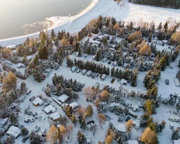 Wintercamping im Erzgebirge: Wohnmobile und Wohnwagen stehen im Schnee auf einem Campingplatz am See, umgeben von verschneiten Bäumen.