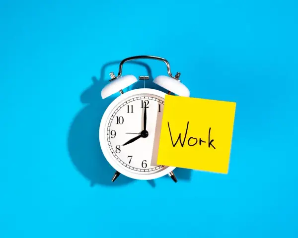 Reloj despertador blanco con nota amarilla que dice "Work", recordando la importancia de la jornada laboral.