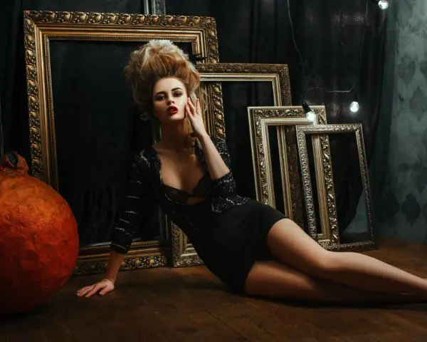 Une femme aux cheveux volumineux, dans une robe noire, pose près de cadres dorés. Un tuto Photoshop pour un look dramatique.