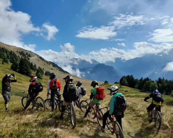 Groupe d'ados en colonie VTT descente, prêts à dévaler les pentes montagneuses sous un ciel bleu éclatant.
