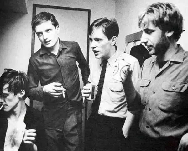 Les membres de Joy Division, Ian Curtis, Bernard Sumner, Peter Hook et Stephen Morris, posent ensemble, certains fumant, d'autres tenant des boissons.