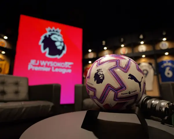 Piłka do gry w Premier League na tle ekranu z logo ligi. W tle widoczne koszulki piłkarskie.