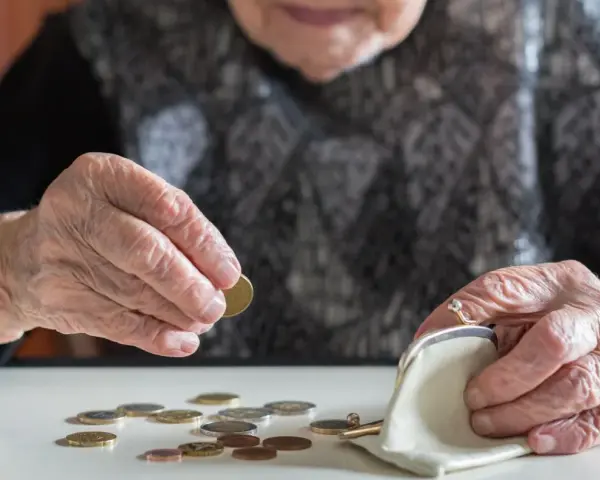 Dłoń starszej kobiety z pierścionkiem wkłada monetę do portmonetki. Rozrzucone monety na stole sugerują trudności finansowe, być może związane z emeryturą po mężu.