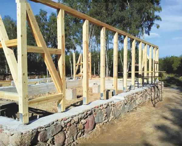 Drewniana konstrukcja domu powstaje na solidnym fundamencie z kamienia. Widać betonowe słupy i kamienny murek.